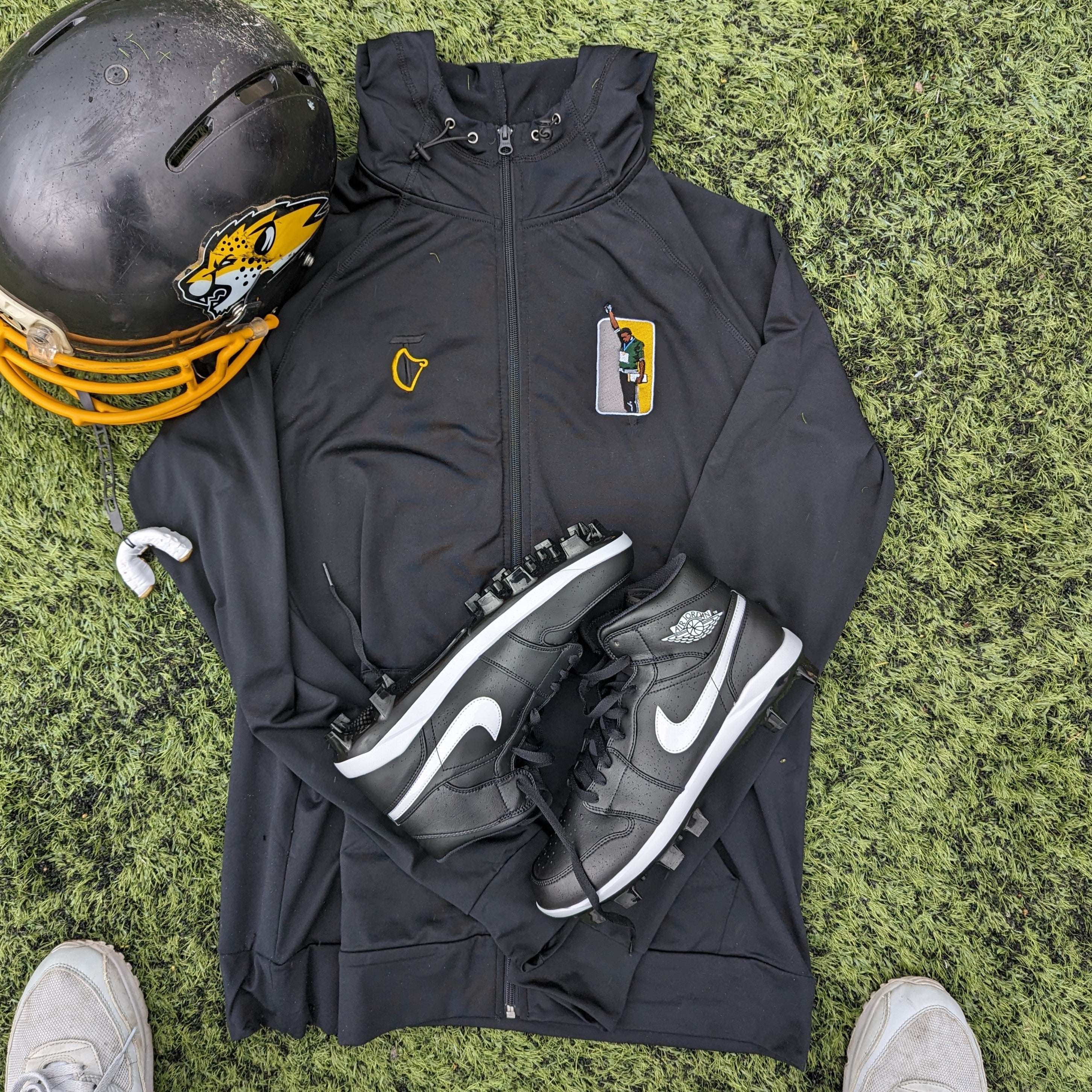 Flatlay image of an athlesuire hoodie with an embroidered design of Tommie Smith raising his black-gloved fists in protest during the 1968 Mexico Olympics Men's 200m medal ceremony. Featuring Jordan 1 Cleats and an American Football helmet with Hertfordshire Cheetah decals