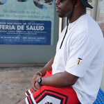 Person wearing a white shirt with a logo and red shorts with a colorful design, standing near a poster.