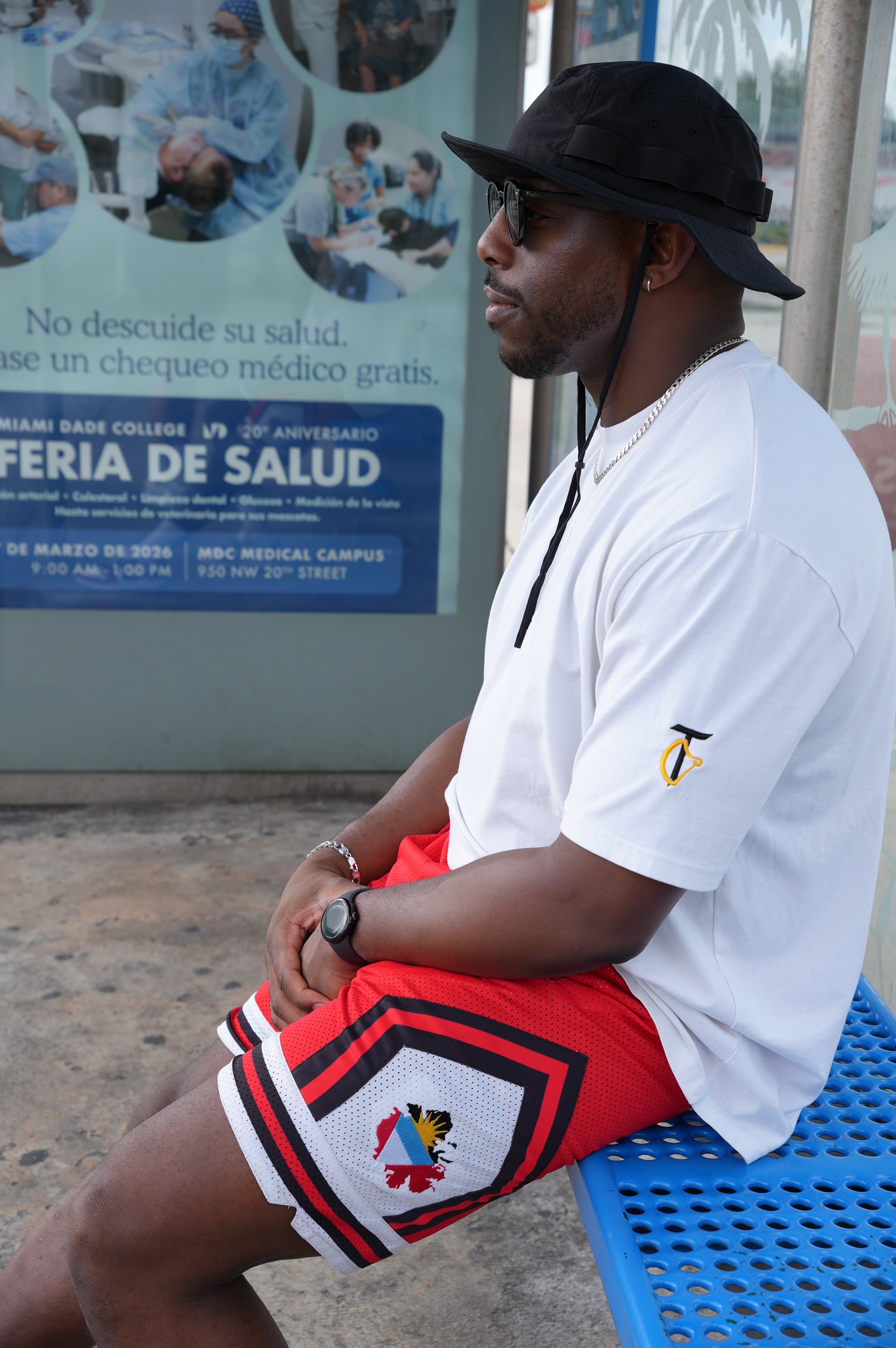 Person wearing a white shirt with a logo and red shorts with a colorful design, standing near a poster.