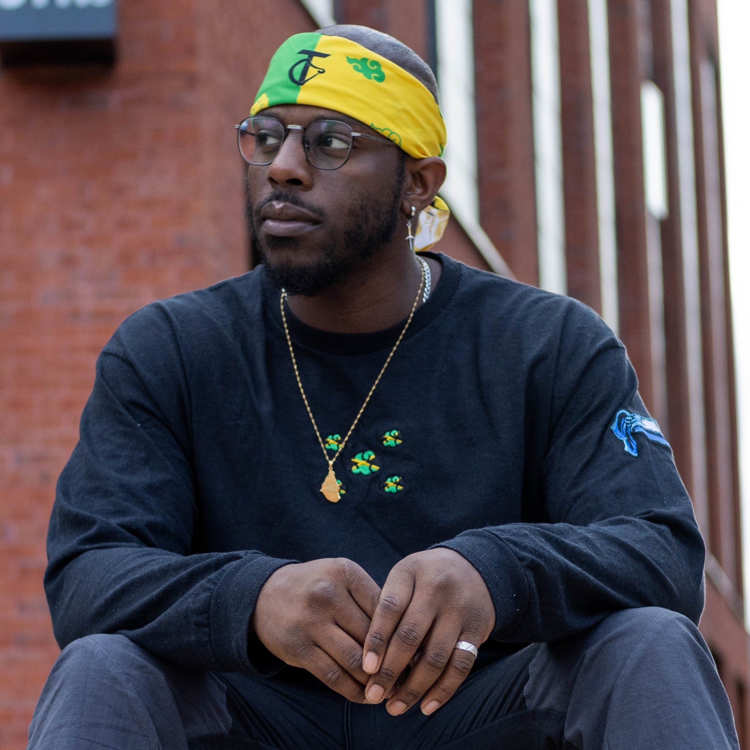Man wearing a black Jamaica Cloud Long sleeve and  green and yellow headband sitting against a brick wall.